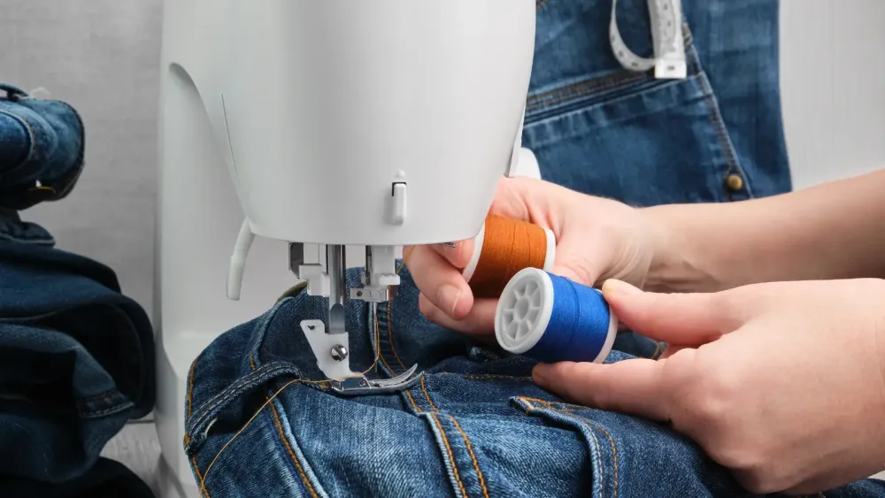 Jeans under a sewing machine beside orange and blue thread
